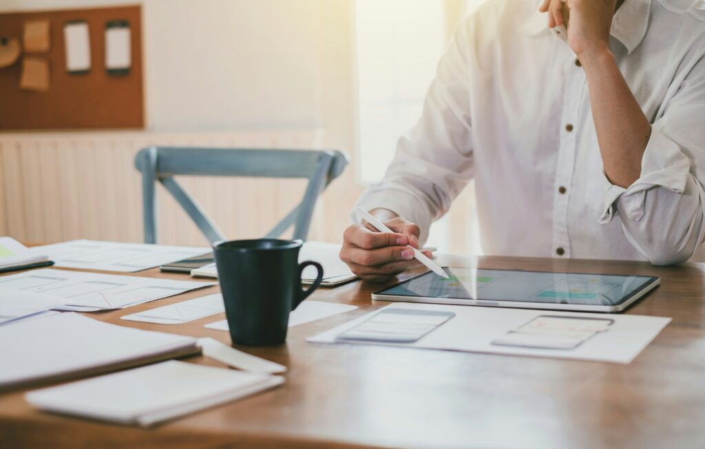 A person working with digital design tools at a wooden table, surrounded by papers, notebooks, and a cup, with natural light in the background.